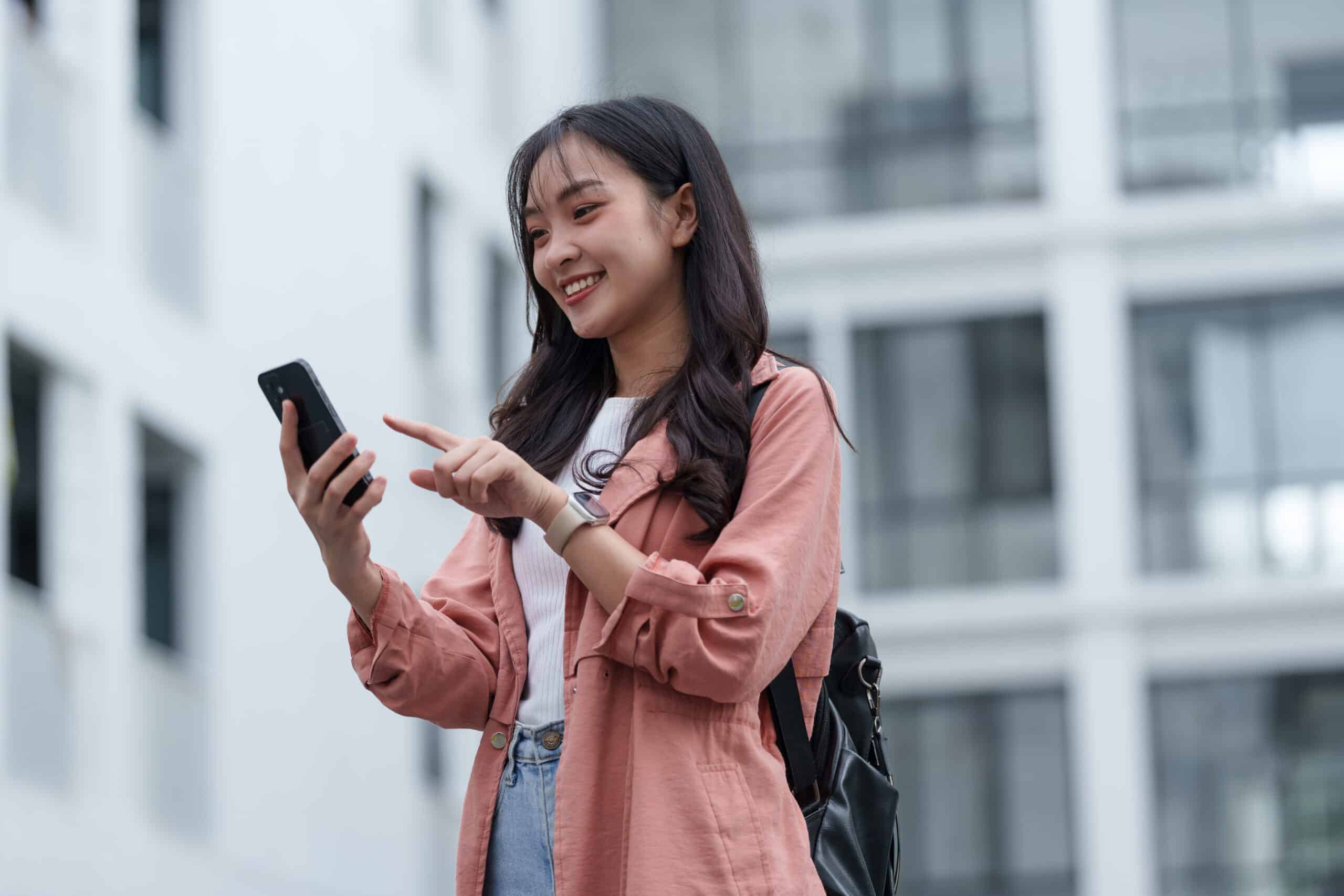 Happy university student using mobile phone app, browsing internet, ordering taxi, booking tickets online, reading message, checking social media, wearing smartwatch and carrying backpack